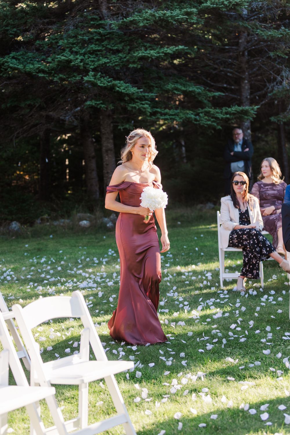Outdoor wedding ceremony setup with white chairs and scattered flower petals on a sunny lawn surrounded by trees.