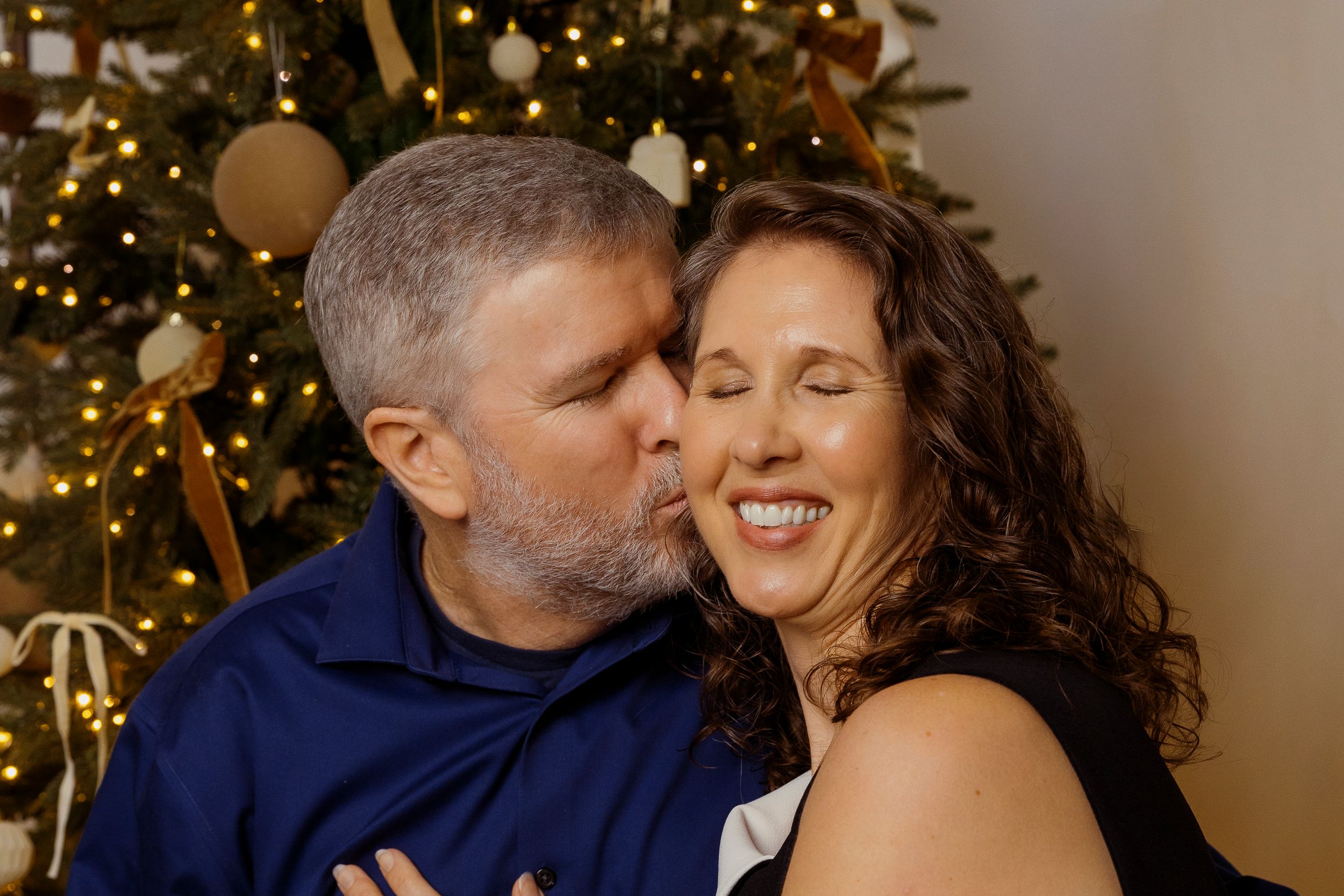 A couple shares an intimate moment in front of a Christmas tree with twinkling lights in the background.