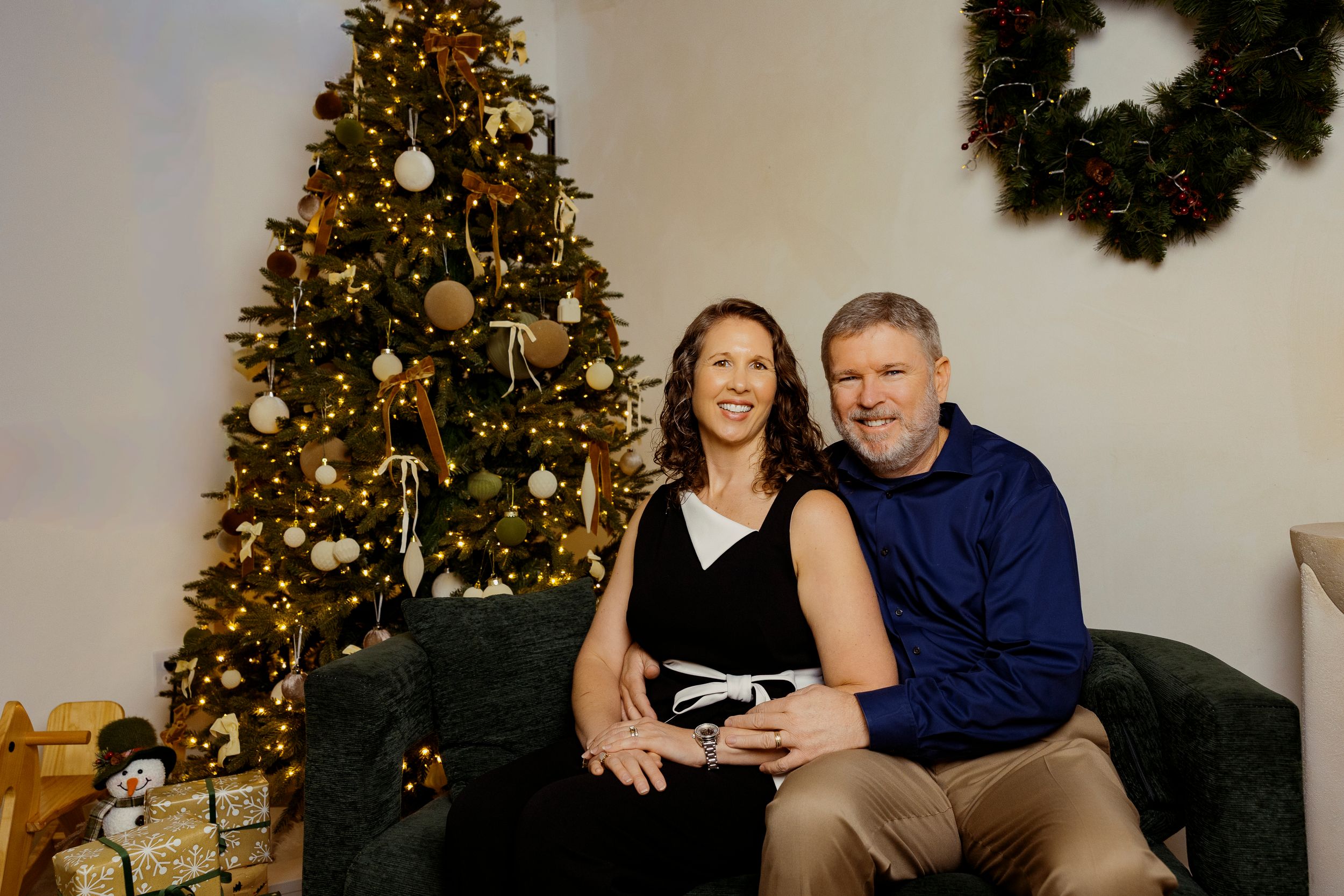 A couple sits together on a couch during a holiday photoshoot with decorated Christmas trees and wreaths in the background.