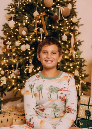 A child wearing festive holiday pajamas sits in front of a beautifully decorated Christmas tree with twinkling lights.