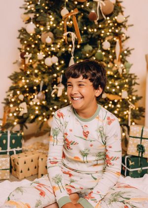Children wearing matching holiday pajamas sit and smile in front of a decorated Christmas tree.