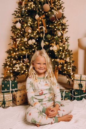 Children in festive white pajamas with holiday prints pose by a lit Christmas tree with wrapped presents.