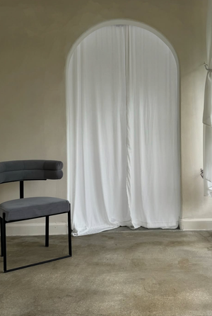 Minimalist interior with an arched doorway covered by white curtains and a modern gray accent chair on concrete flooring.