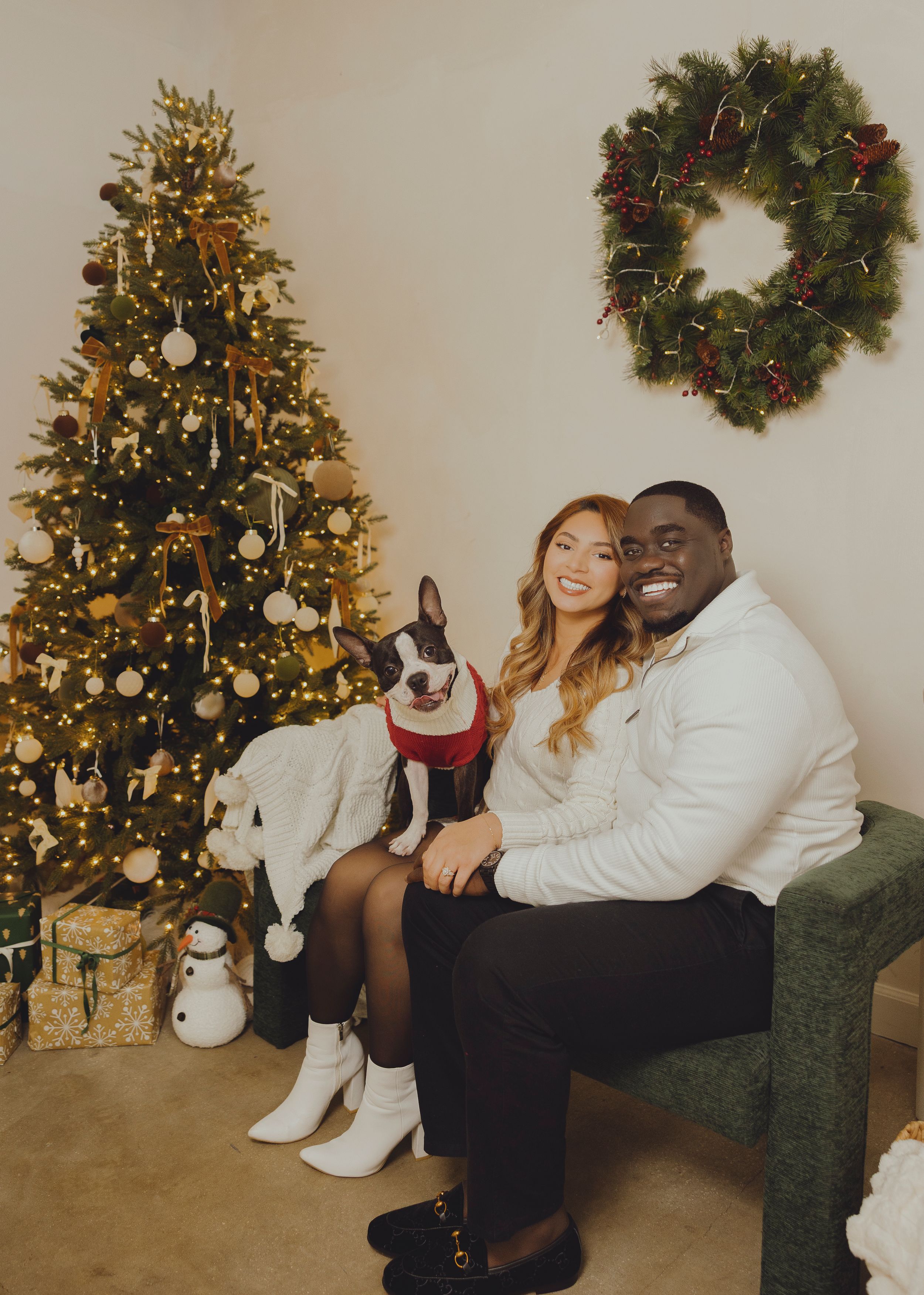 A couple poses together for Christmas photos near a decorated tree and holiday wreath in a cozy home setting.