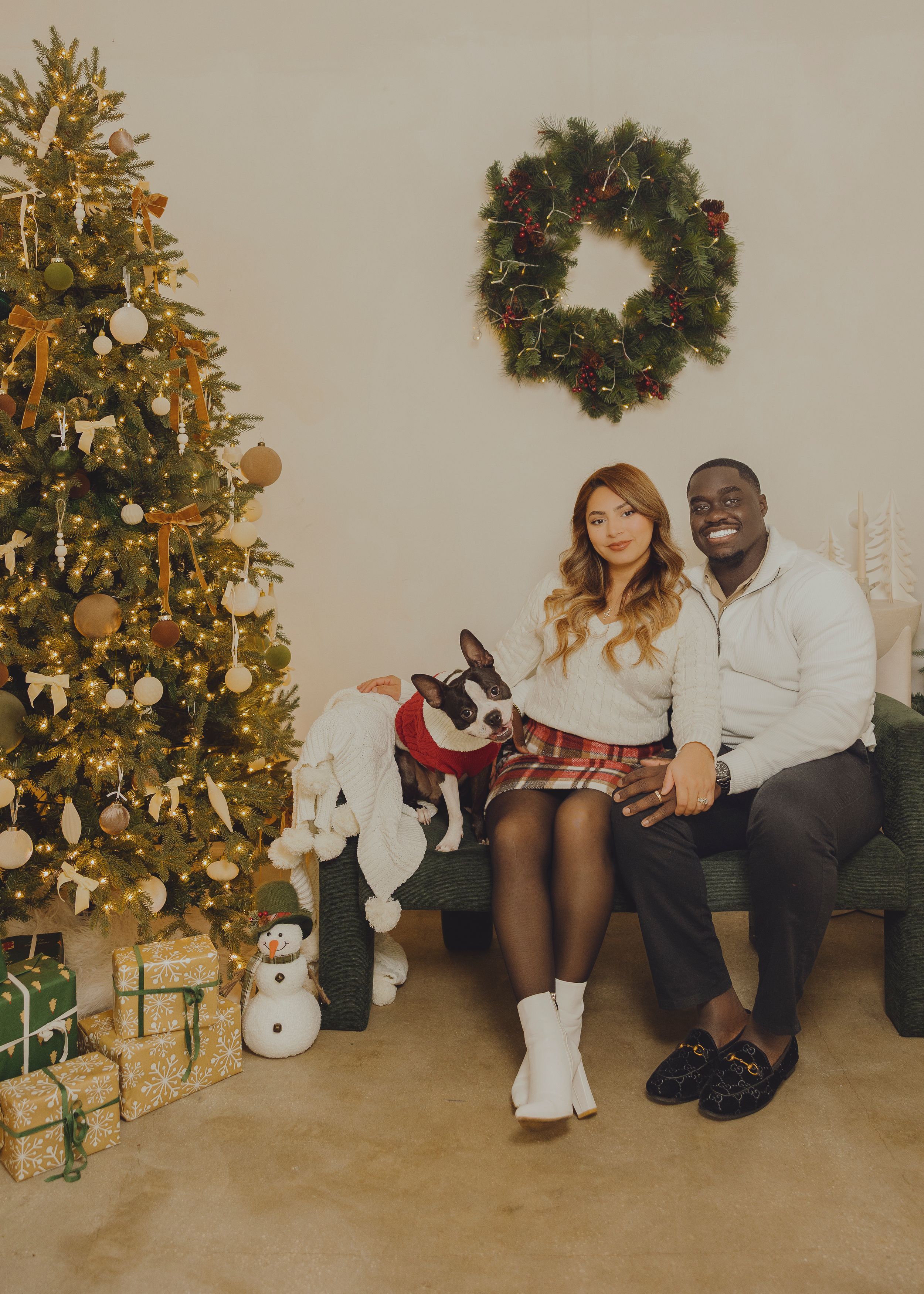A couple sits together on the floor in coordinated white outfits next to a decorated Christmas tree in a holiday photoshoot.