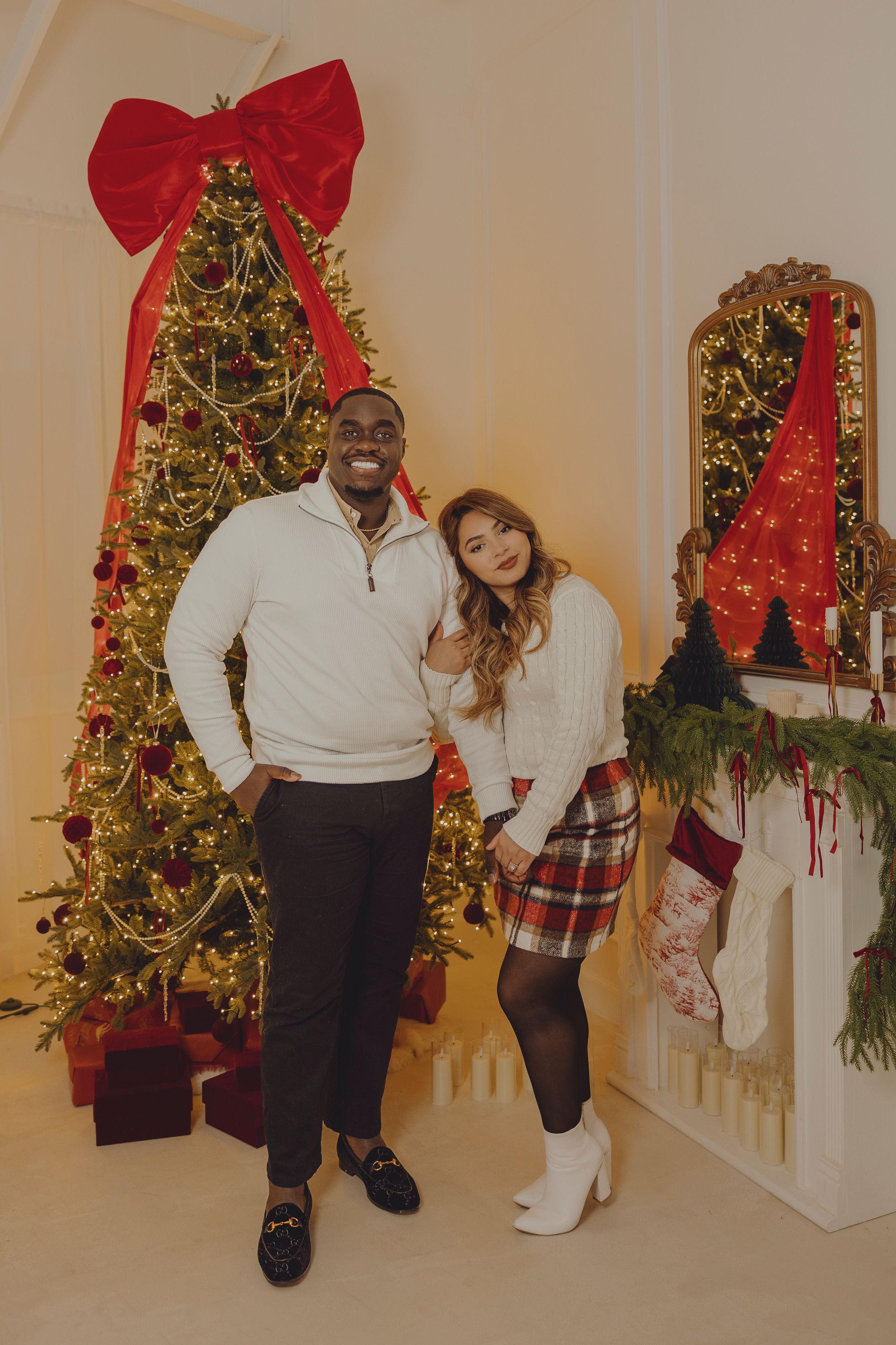 A couple poses in front of a decorated Christmas tree with a large red bow, wearing coordinated white sweaters.