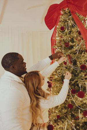 A couple decorates a Christmas tree together, reaching up to hang ornaments on the glowing branches.