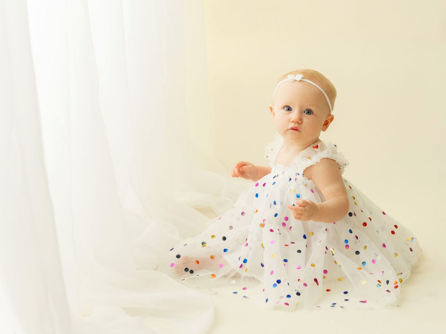 Small baby in white polka dot dress sits near sheer curtains in bright natural light.
