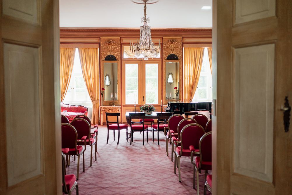 Elegant banquet hall with red velvet chairs and cream walls features multiple crystal chandeliers and large windows.