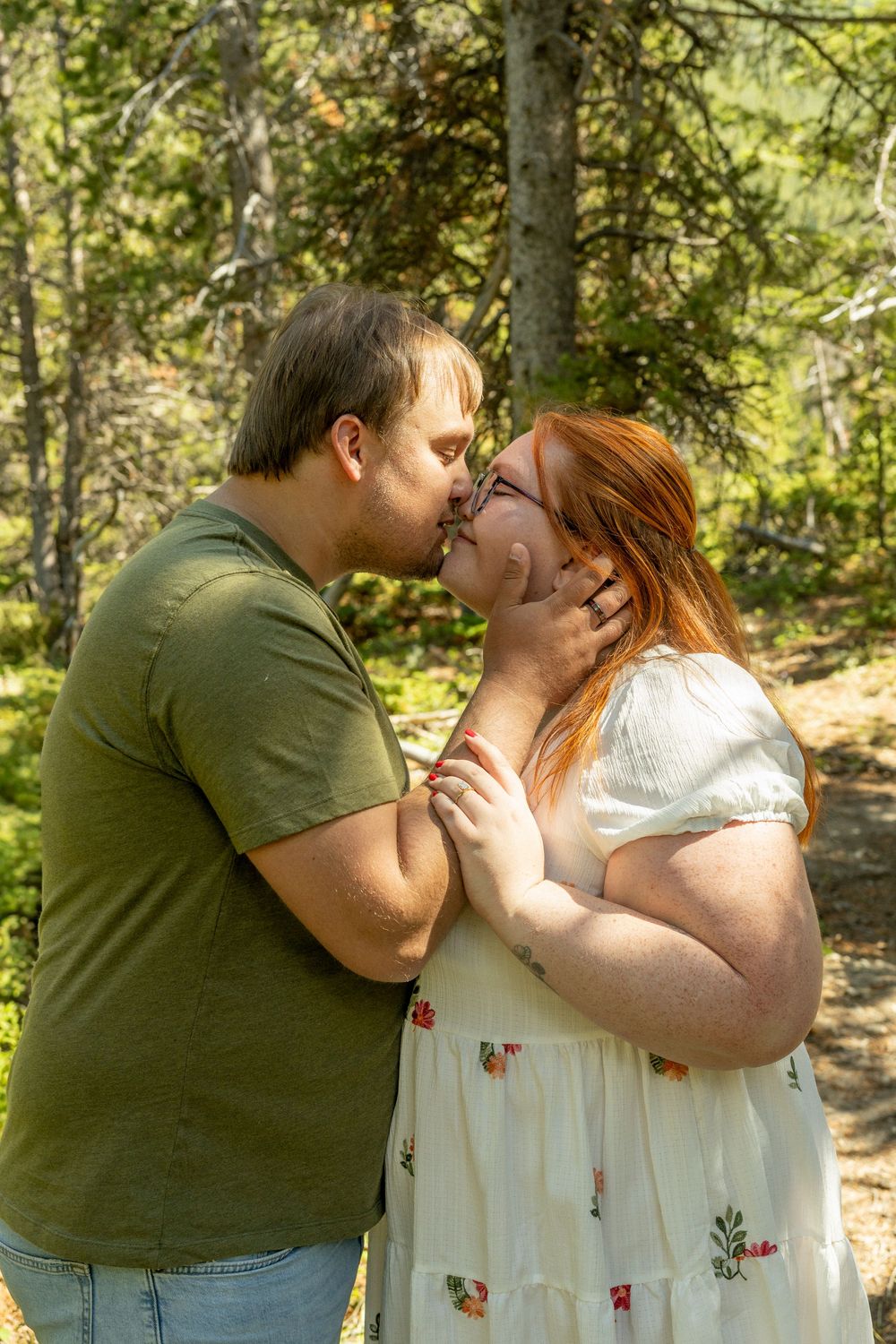 A couple shares a romantic kiss outdoors in a forest setting, with the woman in a floral dress and man in a green shirt.