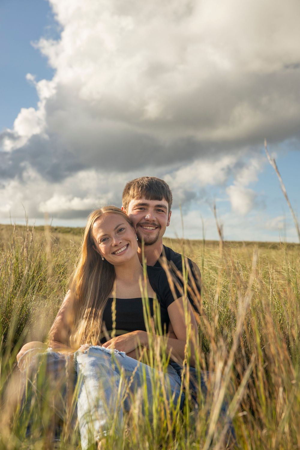 A couple sits together in a golden wheat field under dramatic cloudy skies during sunset.