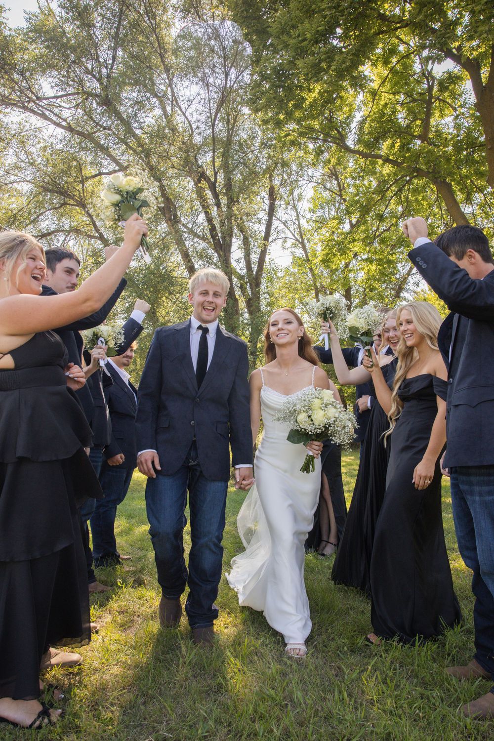 Wedding party celebrating outdoors with bridesmaids in black dresses and newlyweds walking through on grass under trees.
