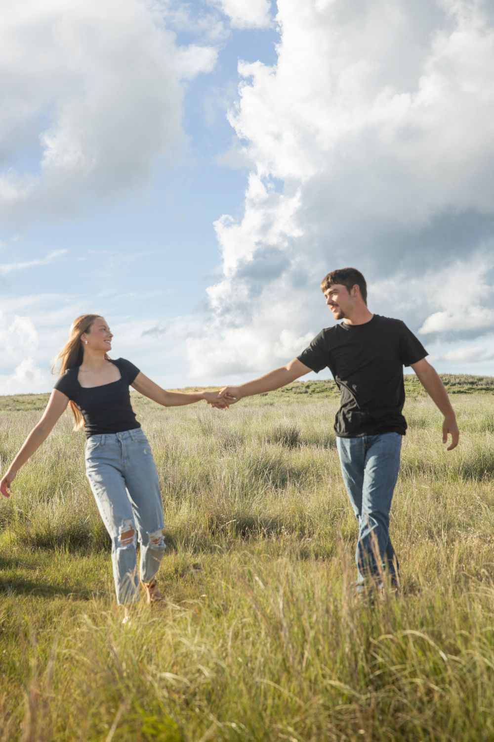 Two people hold hands while walking through a grassy meadow on a cloudy day.