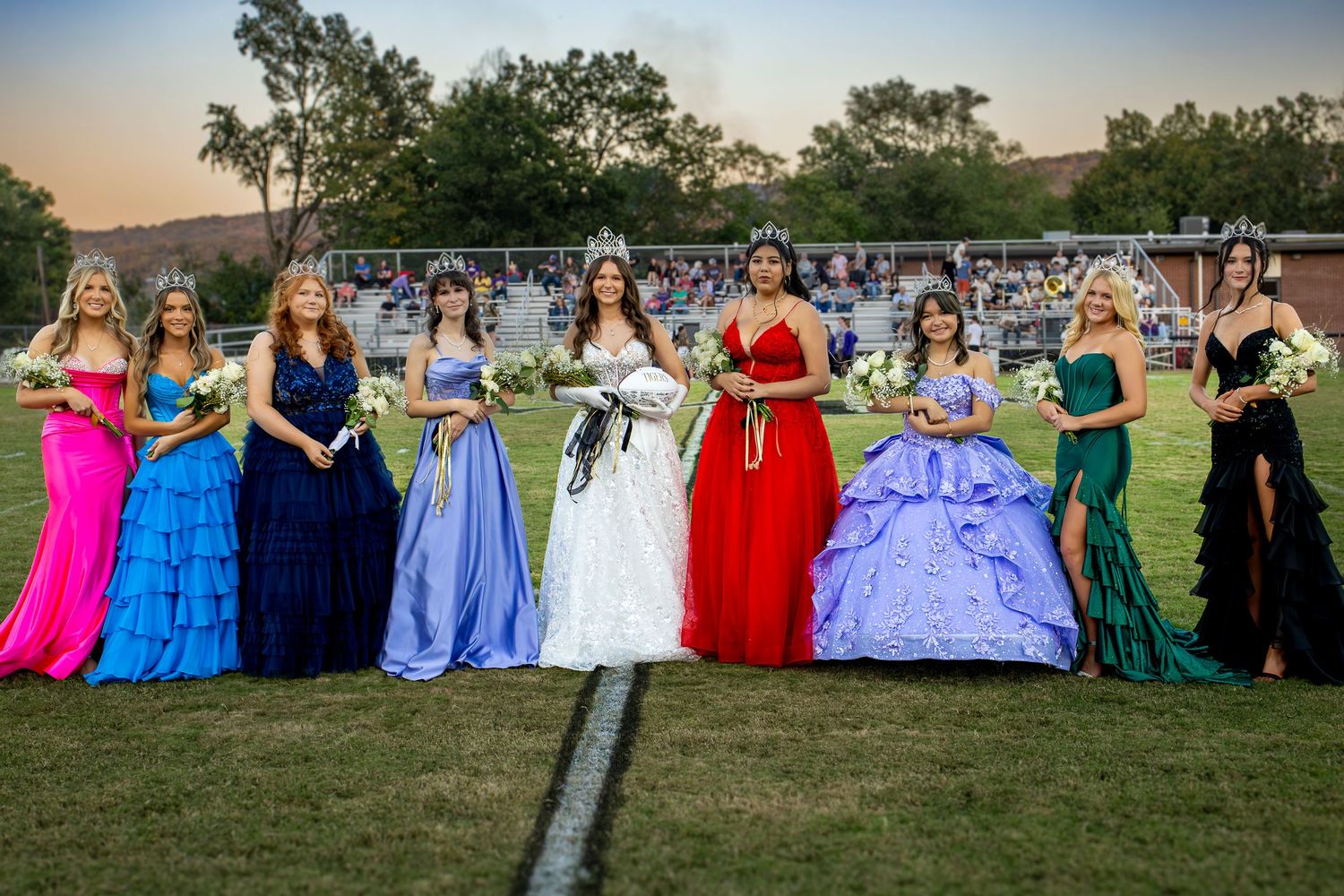 Large group of students in formal attire pose together outdoors for a prom photo at sunset.