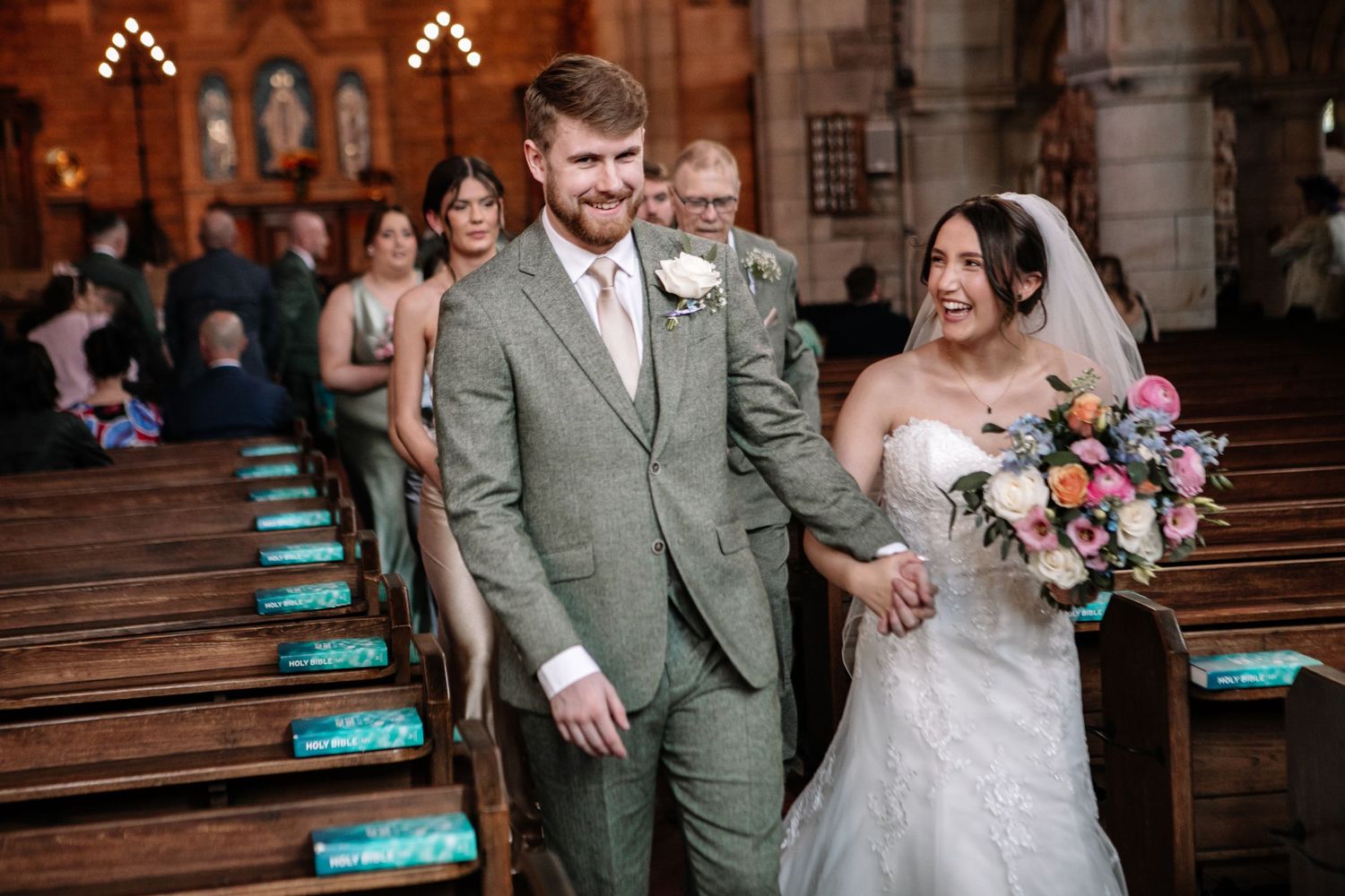 A wedding couple walks down the church aisle together while guests look on from wooden pews with turquoise cushions.