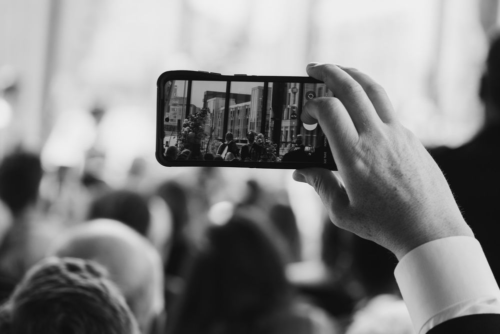 Someone holds up a smartphone to record video of a crowded event in black and white.