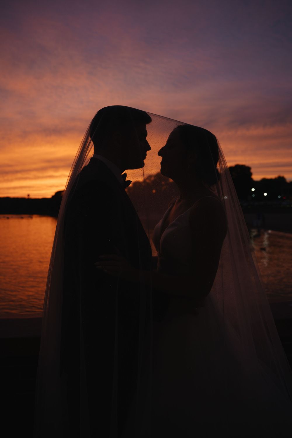 Silhouette of bride and groom under veil at sunset by water, vibrant orange and purple sky backdrop.