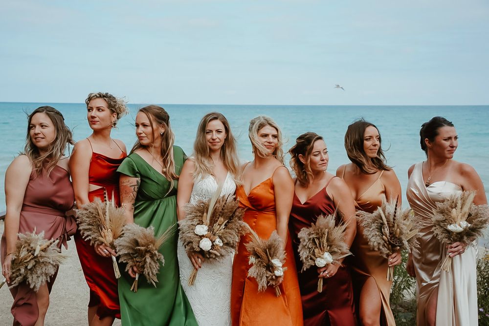 Wedding party members in orange, green, and neutral dresses stand in a line against a bright blue ocean backdrop.
