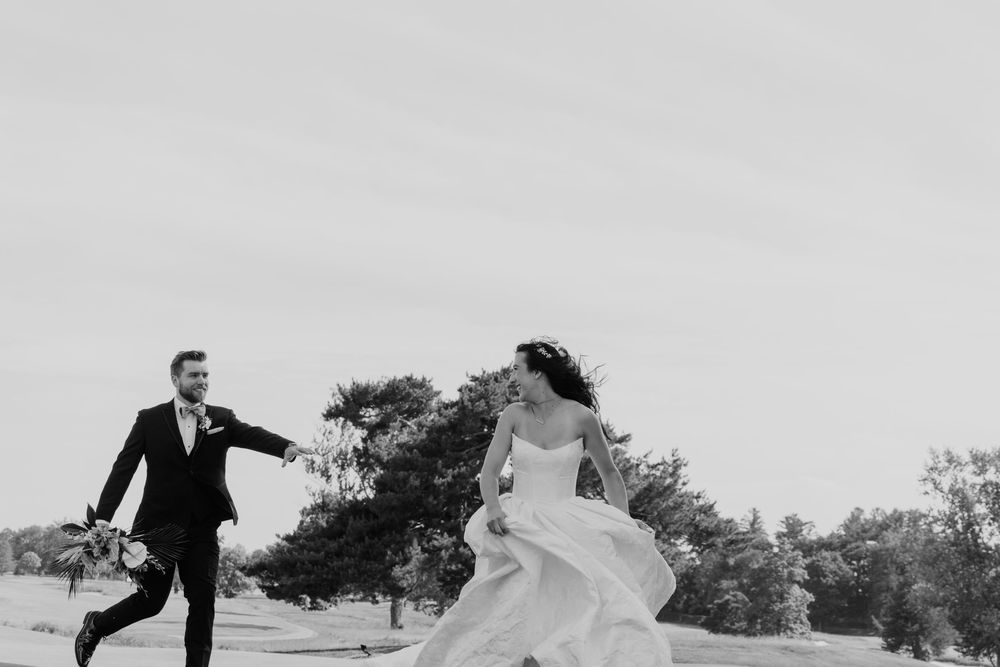 Black and white photo of a bride and groom joyfully running outdoors on their wedding day.