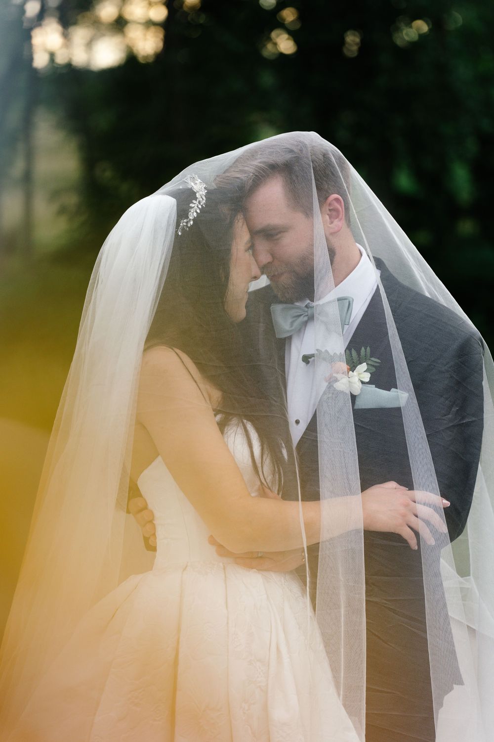 Bride and groom share a romantic moment under a flowing white veil outdoors, with soft golden light in the background.