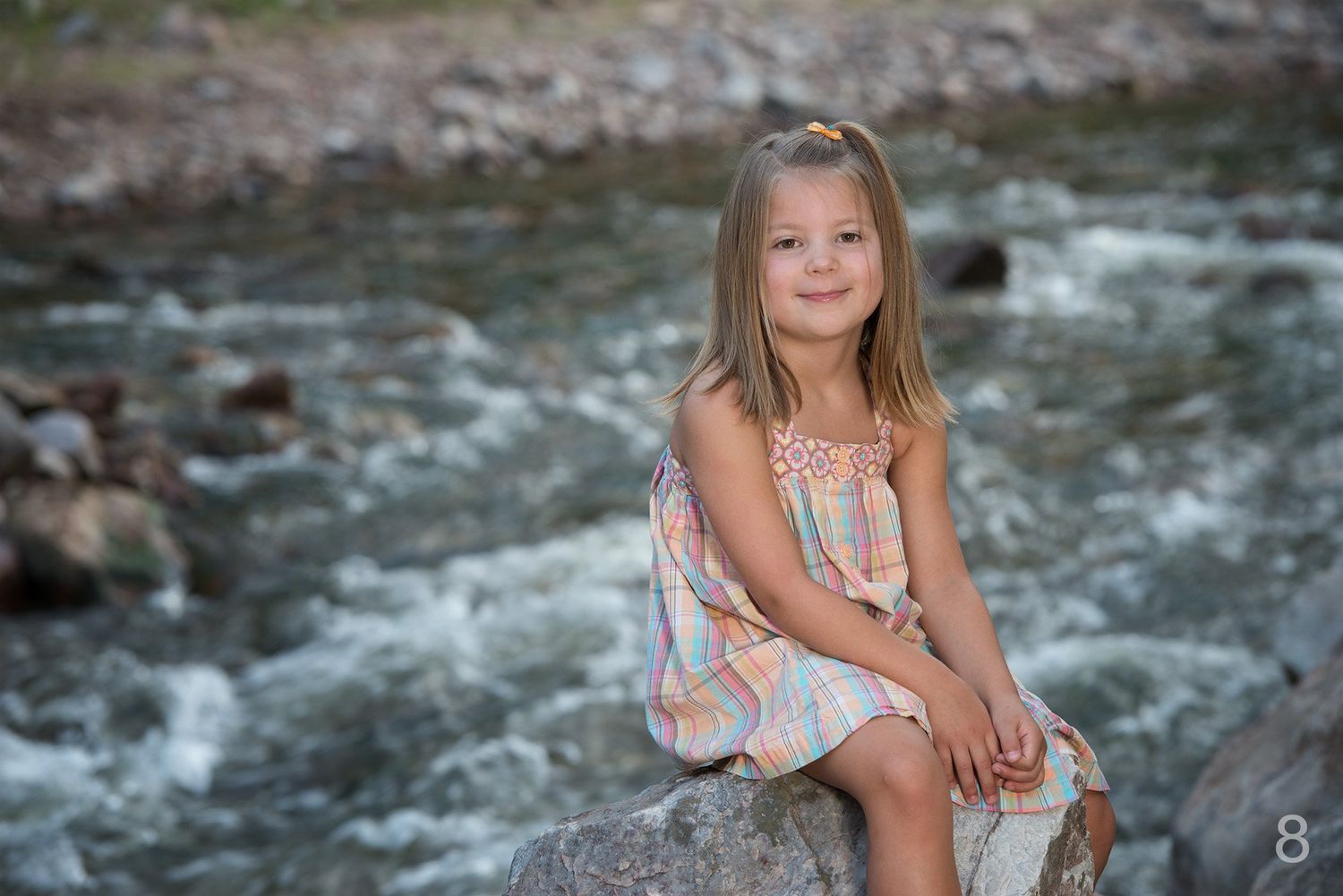 A Erie Colorado child sits along rocky riverbank surrounded by green forest and flowing mountain stream.