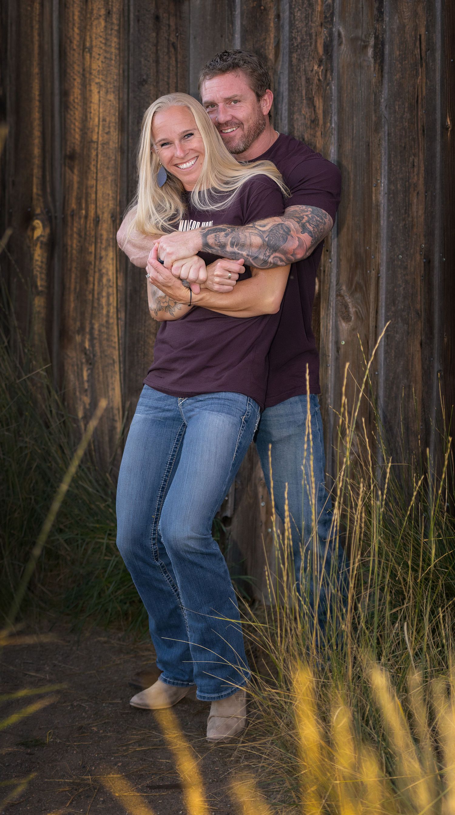 A Thornton Colorado couple embraces and laughs together in front of a rustic wooden fence with tall grass in the foreground.