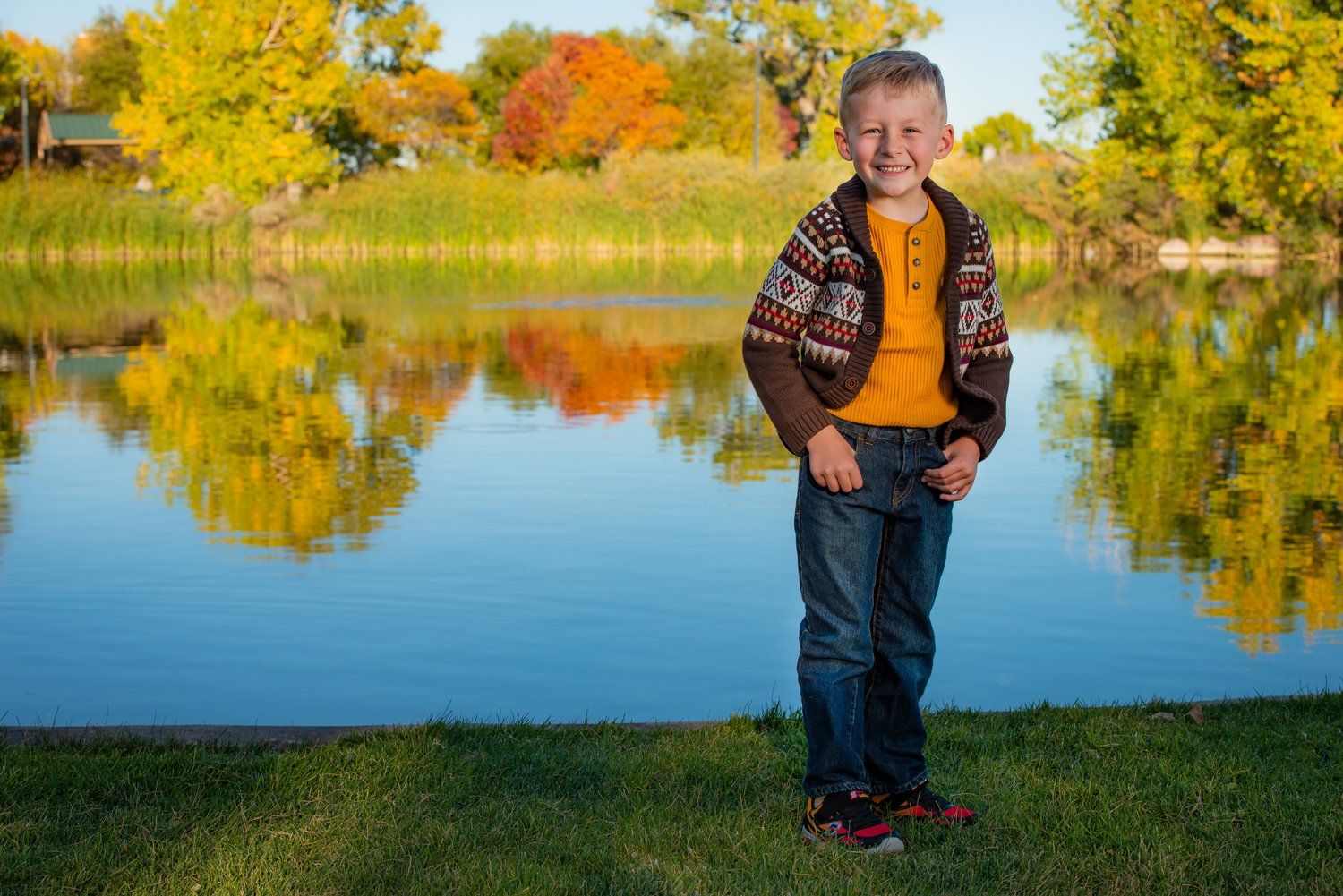 A Thornton Colorado child stands amid autumn landscape with colorful fall foliage reflecting in calm lake water during golden hour.
