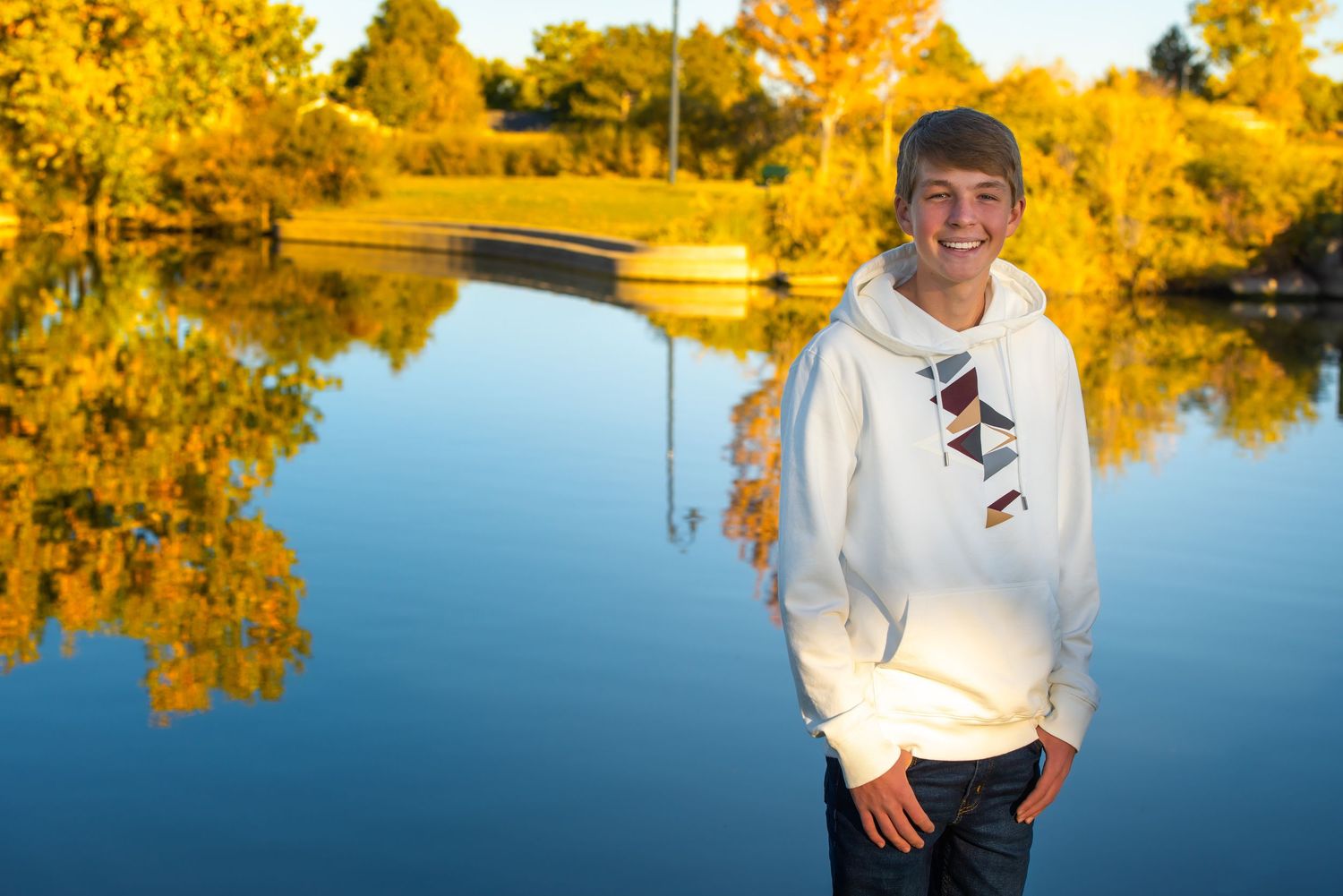 A Thornton Colorado child stands amid autumn landscape with colorful fall foliage reflecting in calm lake water during golden hour.