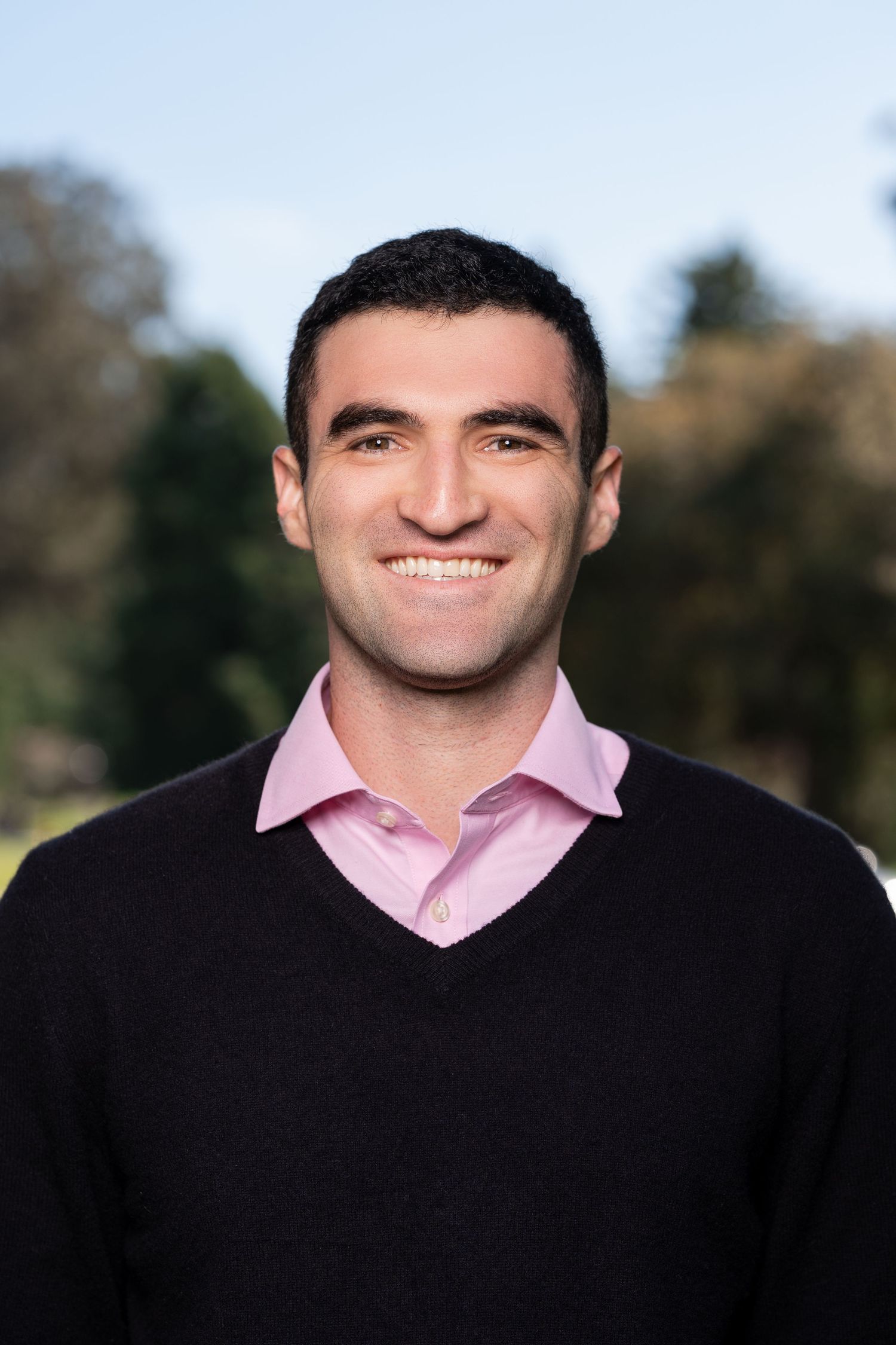 Professional headshot of someone wearing a black blazer and pink dress shirt against a blurred outdoor background.