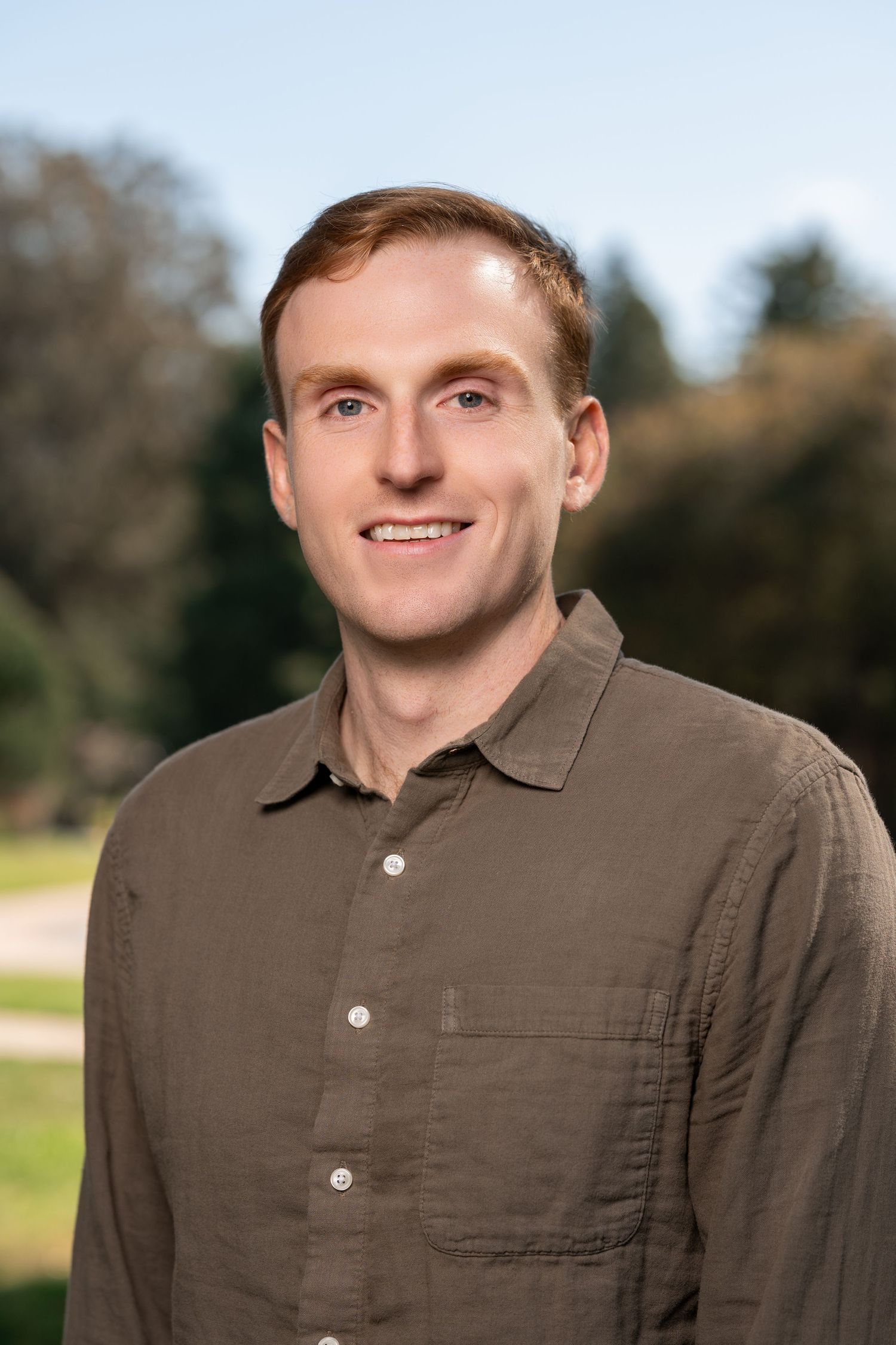A person wearing a brown collared shirt smiles while standing outdoors with trees in the background.