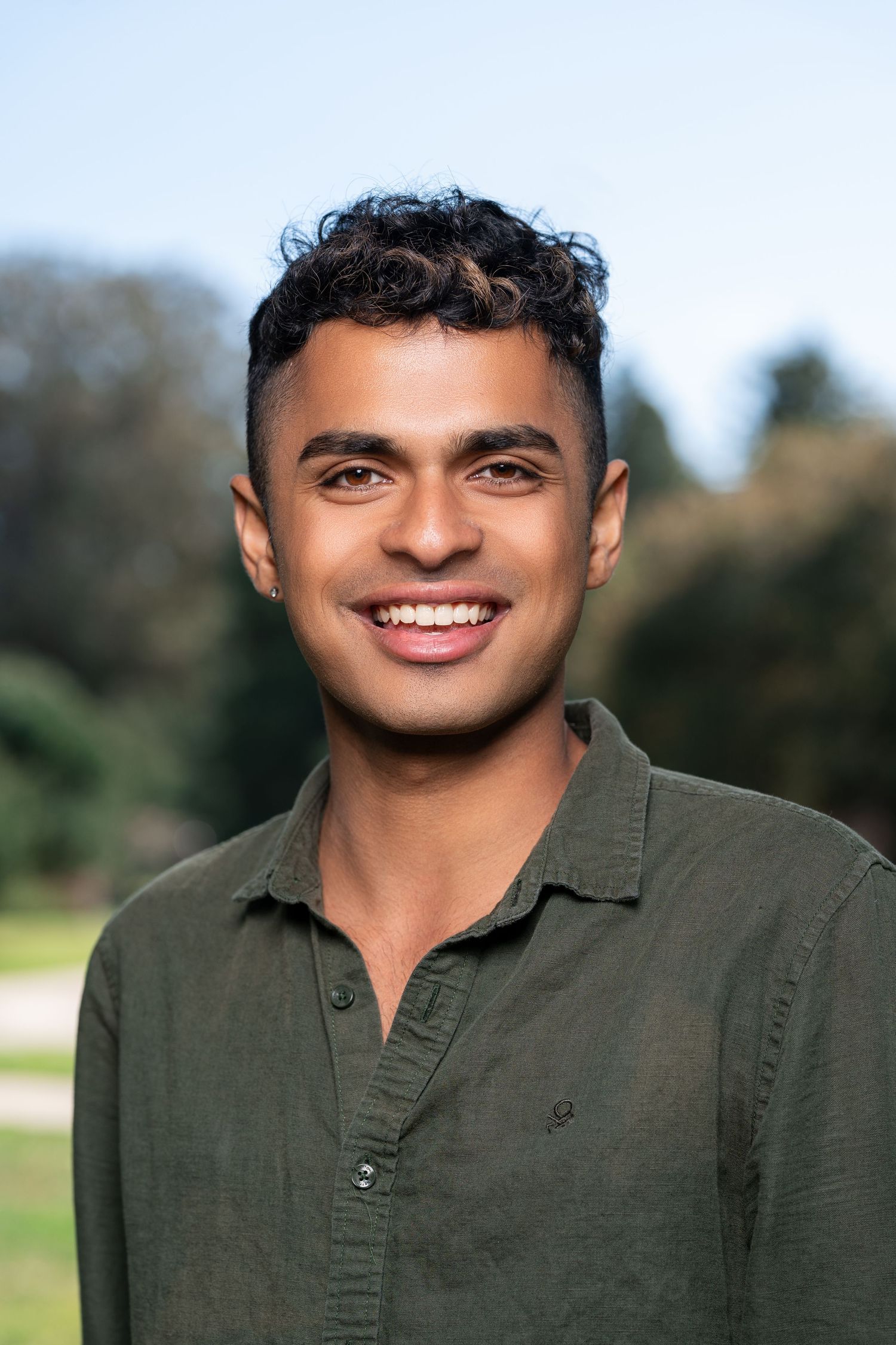 Young person wearing an olive green polo shirt smiles warmly against a blurred outdoor background.