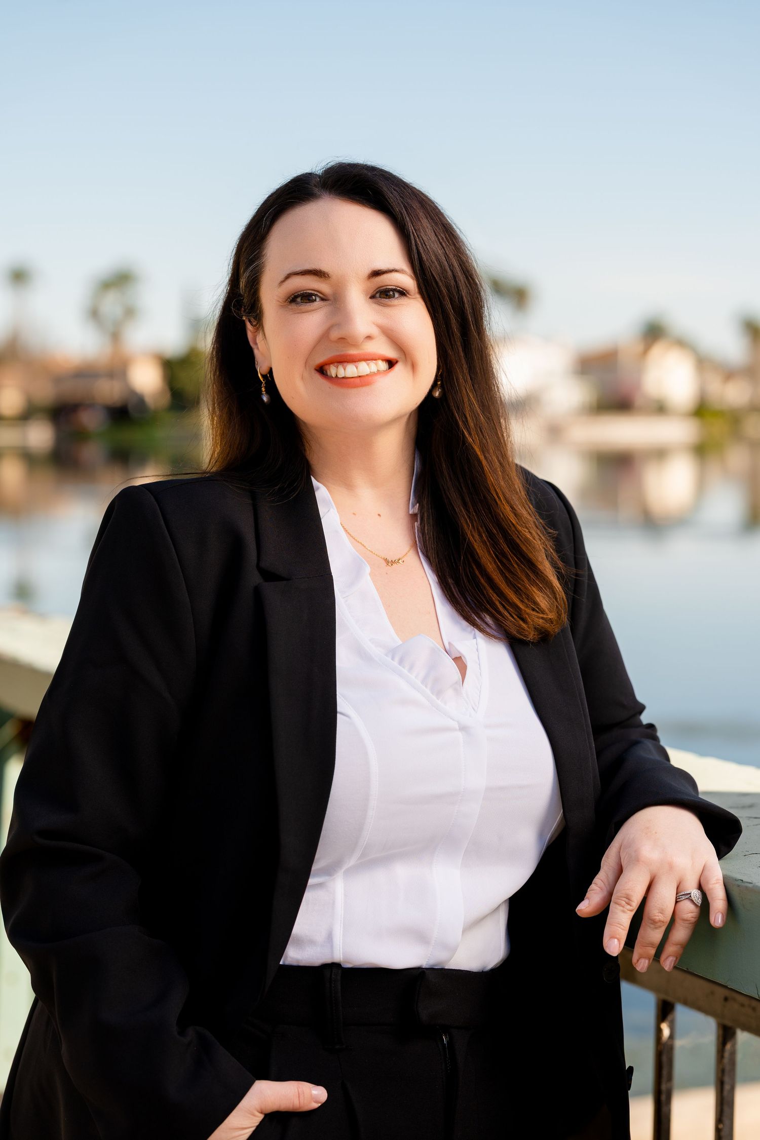 Professional headshots of someone in a black blazer and white blouse smiling by a waterfront setting.