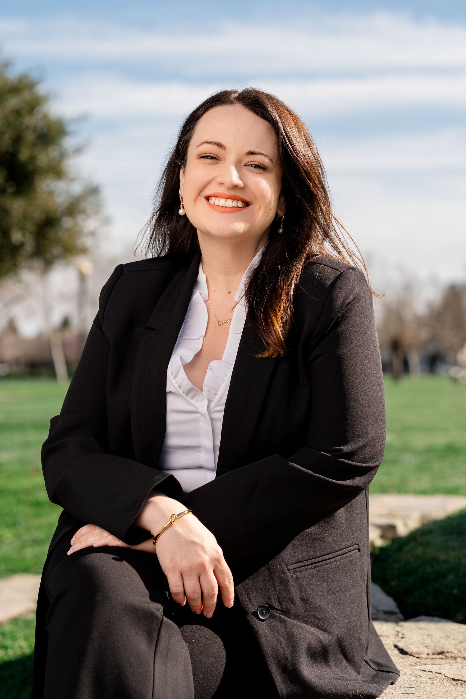 Professional in a black blazer and white shirt sits outdoors on a stone wall against a natural backdrop.