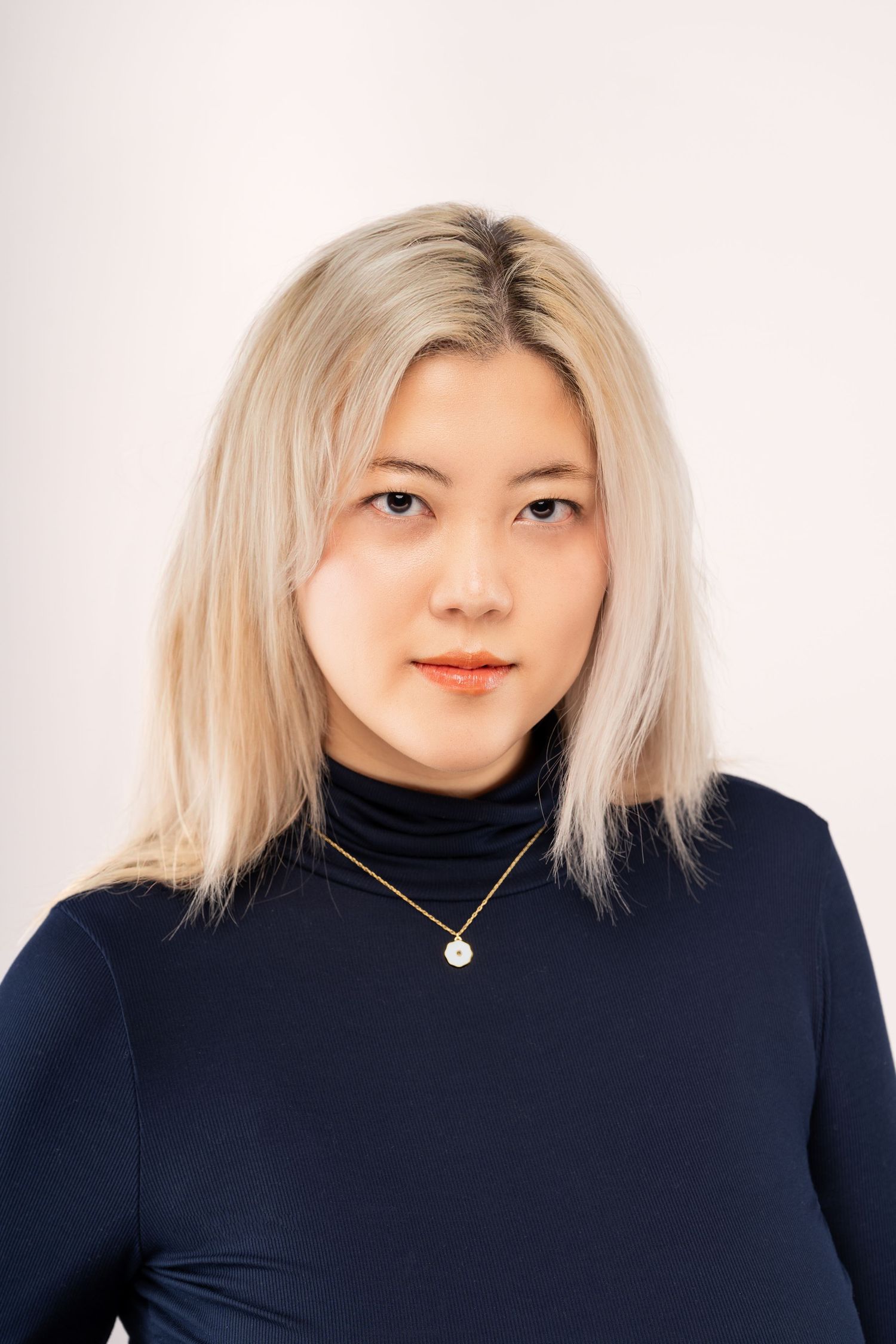 Professional portrait of a person with platinum blonde hair wearing a black turtleneck and delicate necklace against light background.