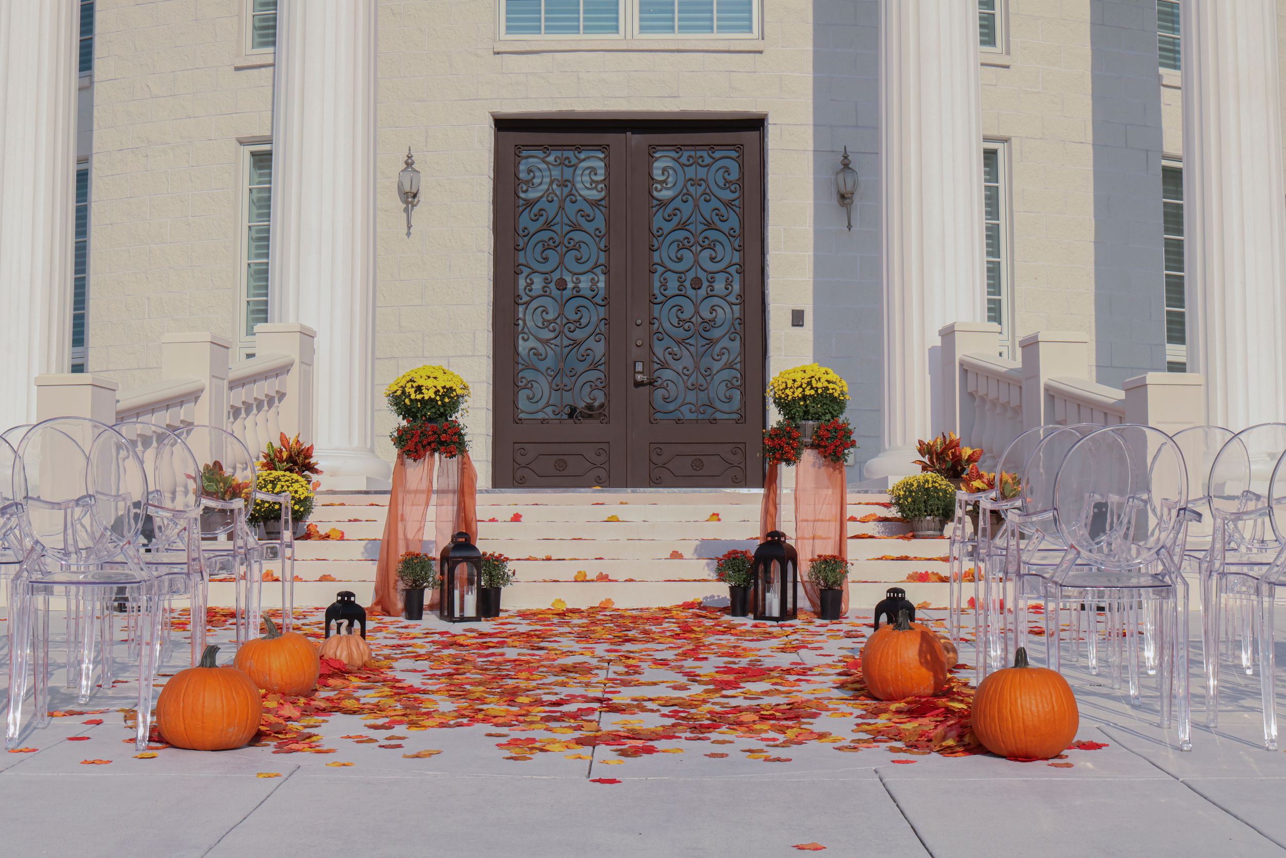 A series of fall decorations with pumpkins and mums arranged along a white church entrance with clear chairs.