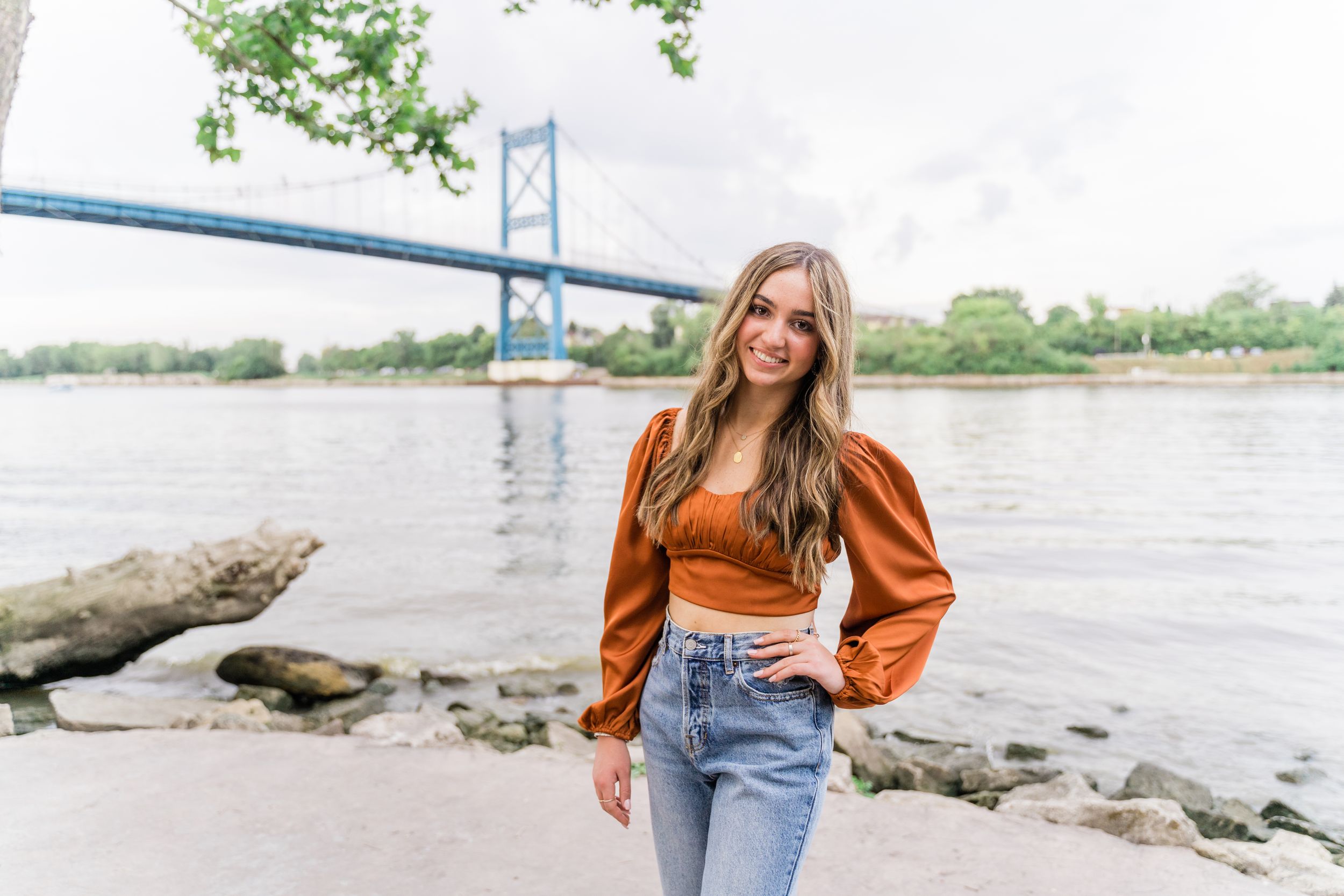 Woman in brown puff sleeve top and jeans poses near waterfront with bridge in background.