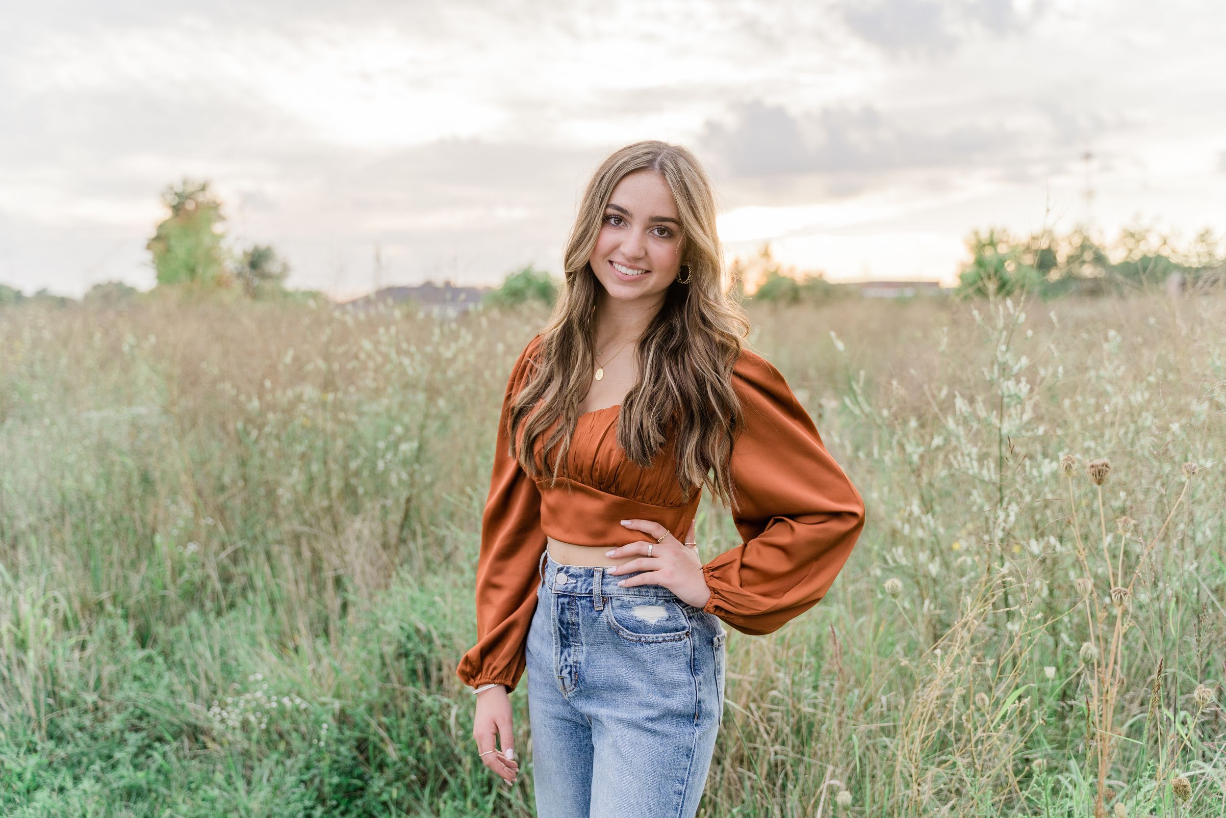 A person in a rust-colored blouse and blue jeans poses in a sunlit field of tall grass and wildflowers.