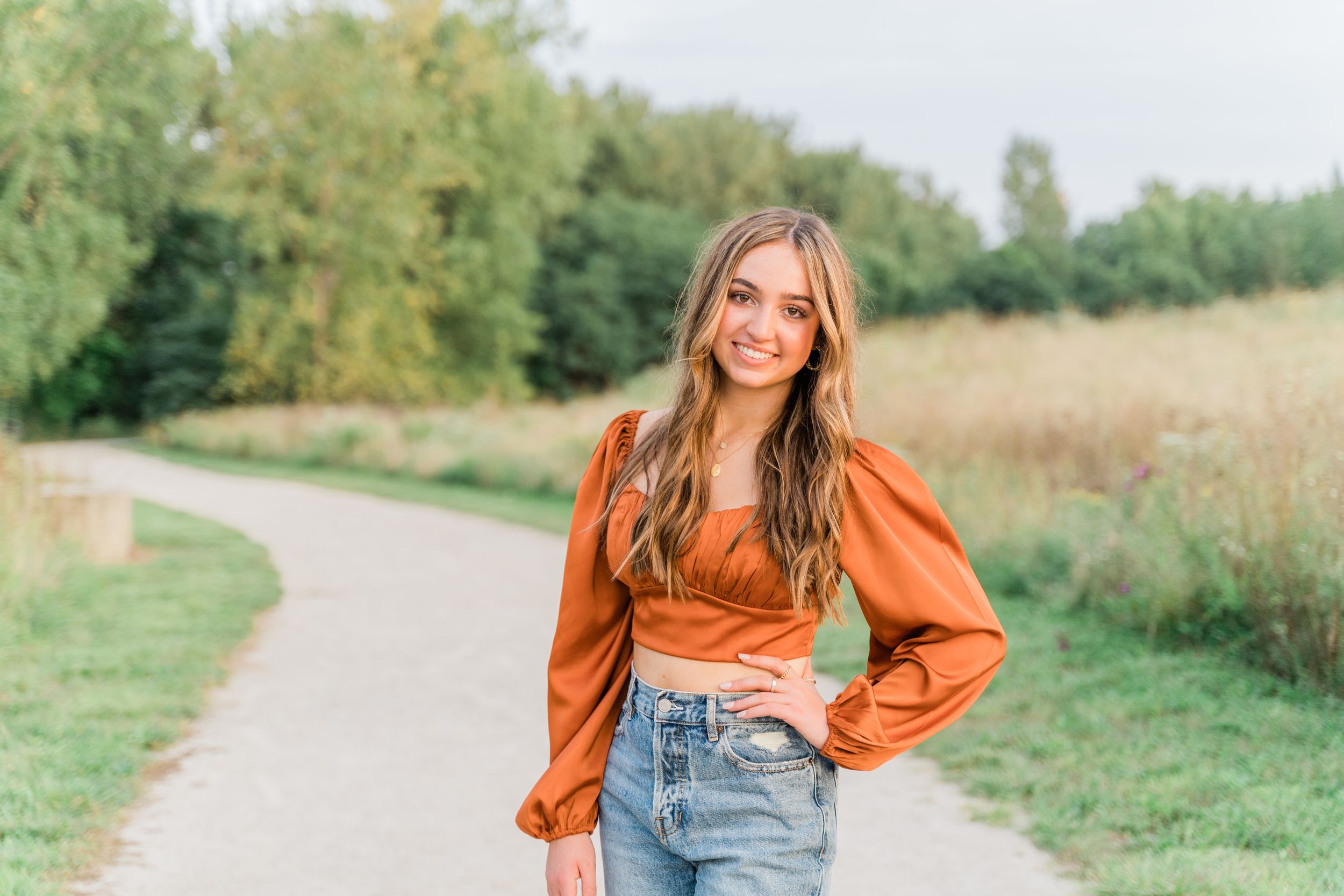 A person in an orange top and denim overalls poses in a sunny meadow with tall grass and trees in the background.
