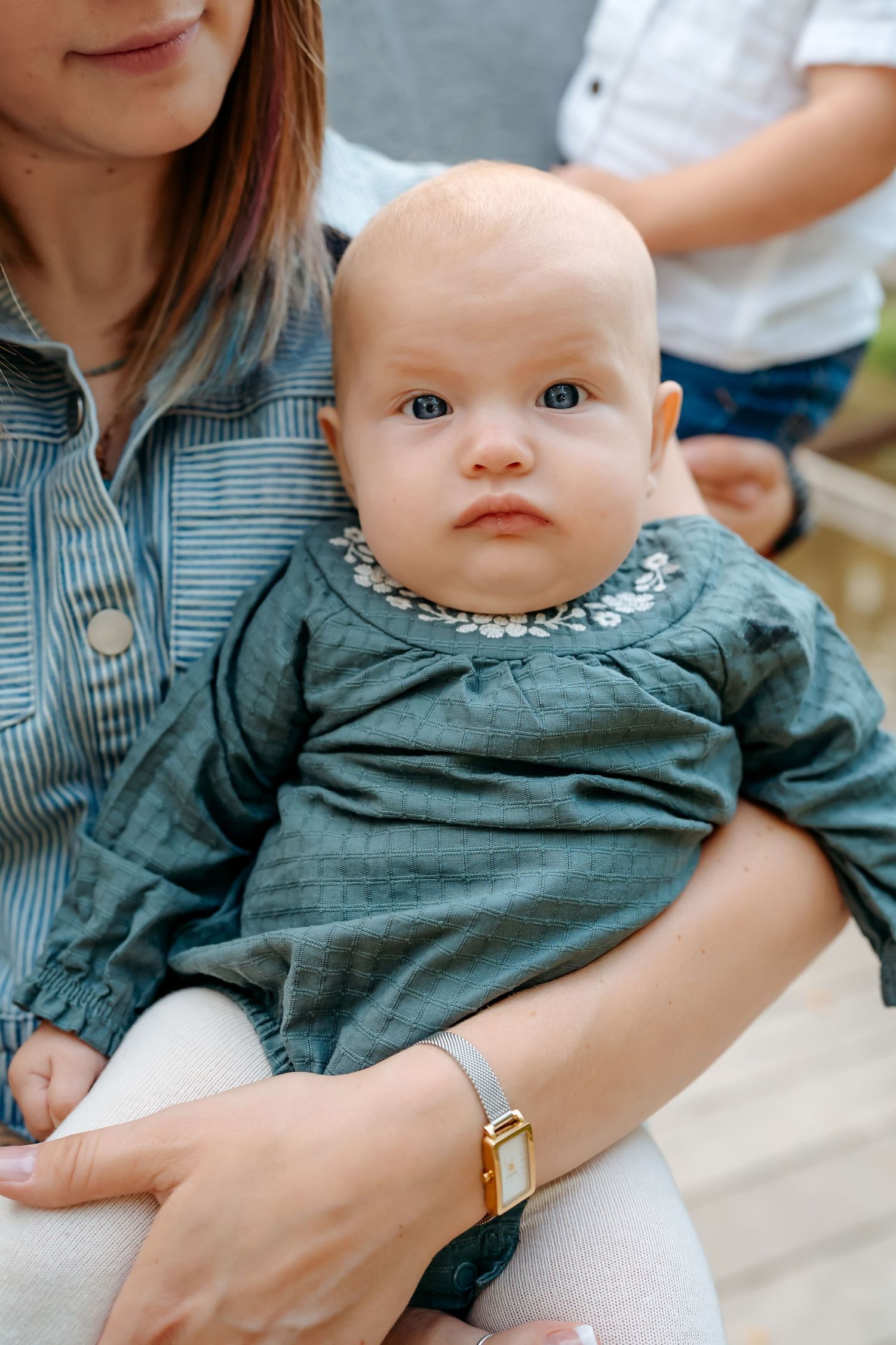 A baby in a teal dress with embroidered collar sits on someone's lap wearing a denim shirt.