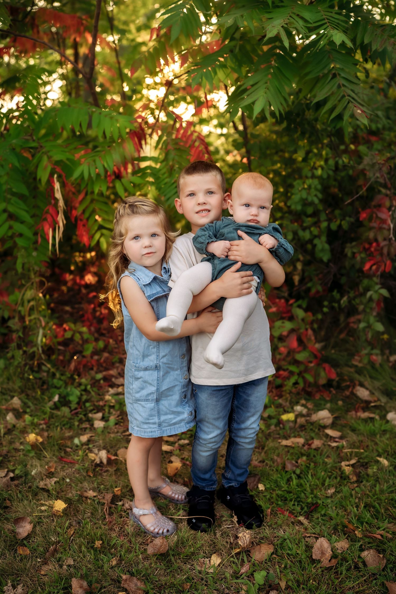 Three children stand together in a garden setting with fall foliage and golden sunlight filtering through trees.