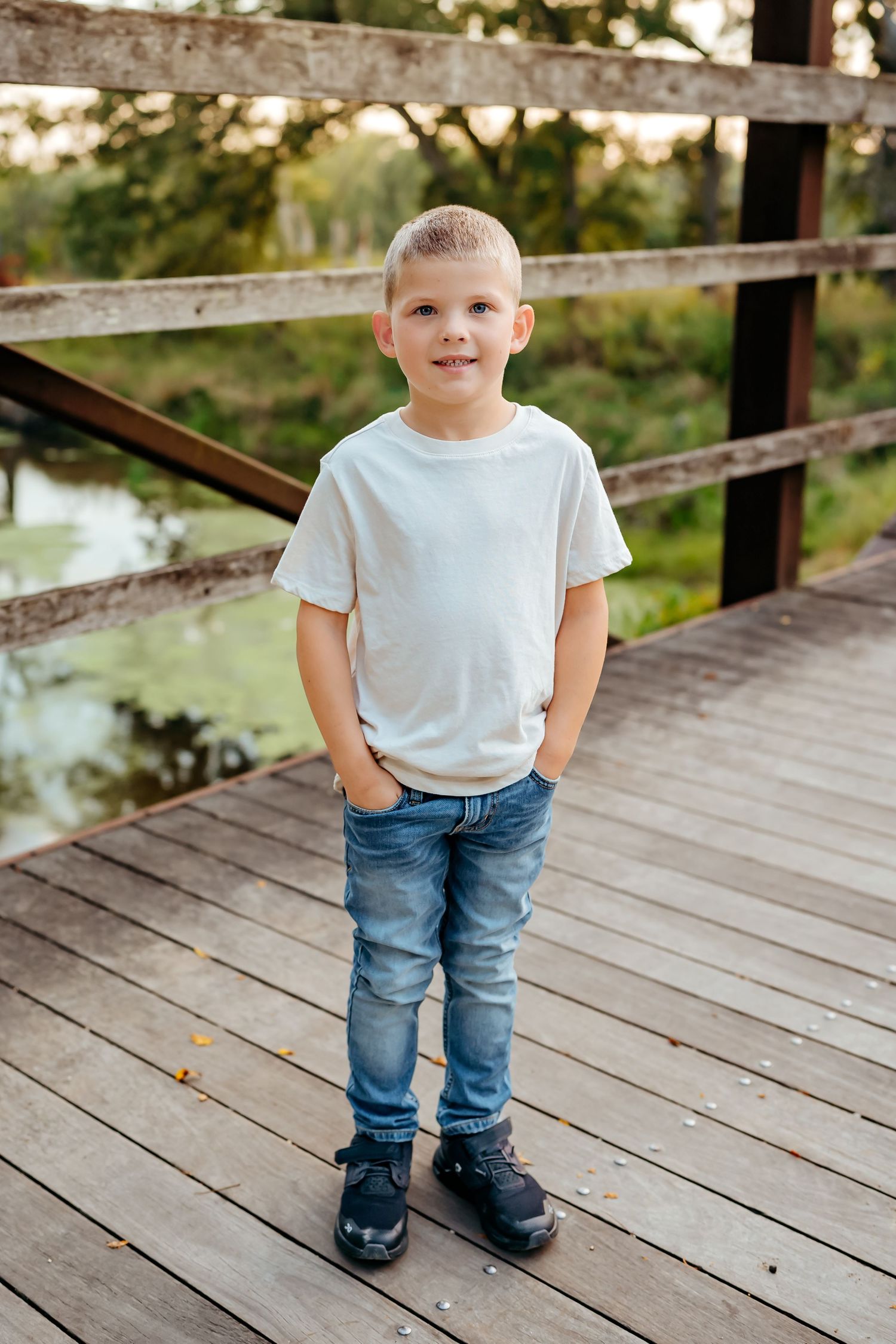 A young child in a white t-shirt and jeans stands on a wooden bridge in an outdoor setting.