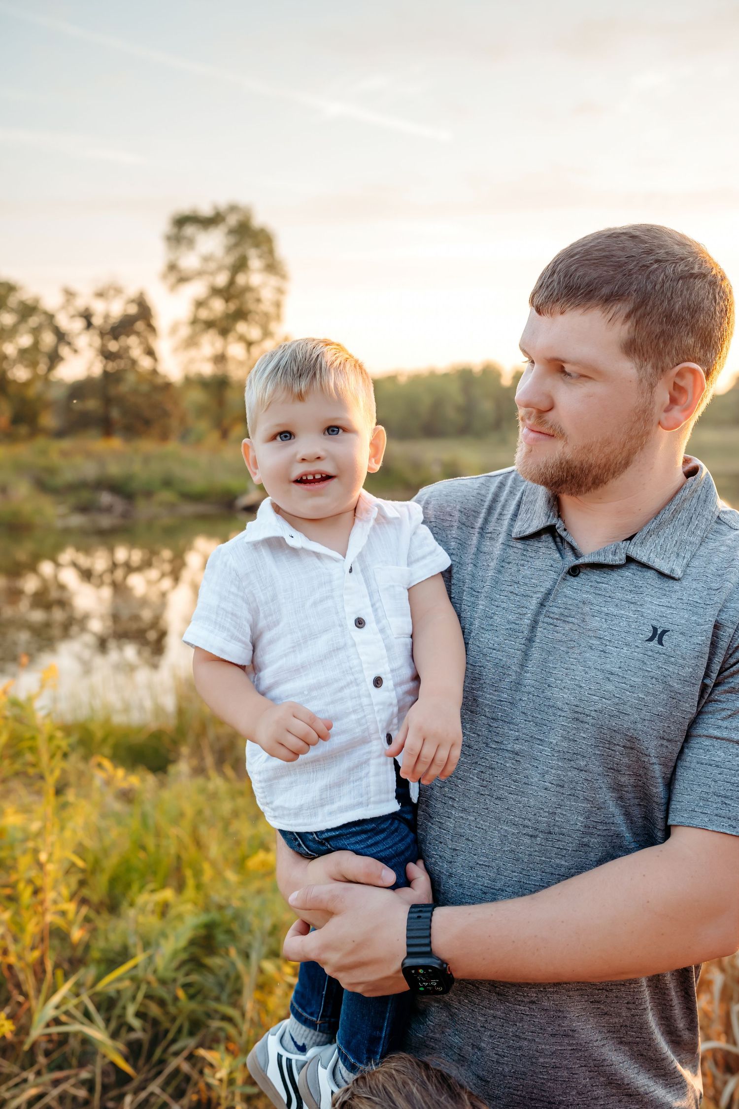 A parent holds a small child in a grey shirt against a golden sunset background in a grassy field.