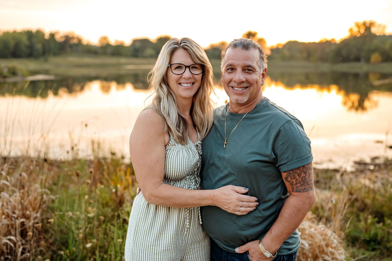 A couple poses together in a field at sunset near a lake with golden light illuminating the natural landscape.
