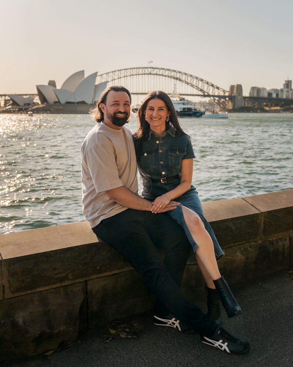 A couple sits together on a waterfront wall with the iconic Sydney Opera House and Harbour Bridge in the background during a photoshoot