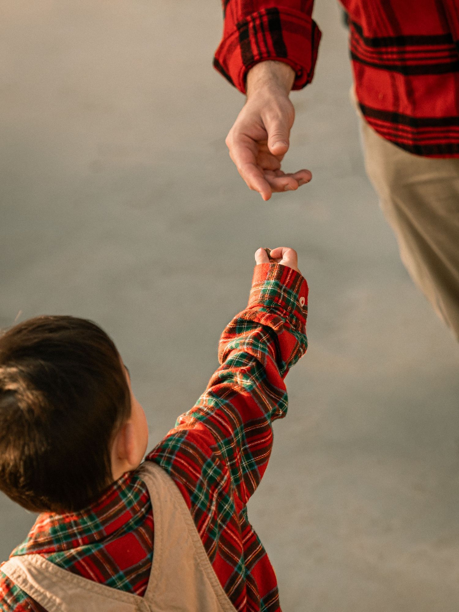 A child in a red plaid shirt reaches up towards someone's hand in a tender moment against a neutral background.