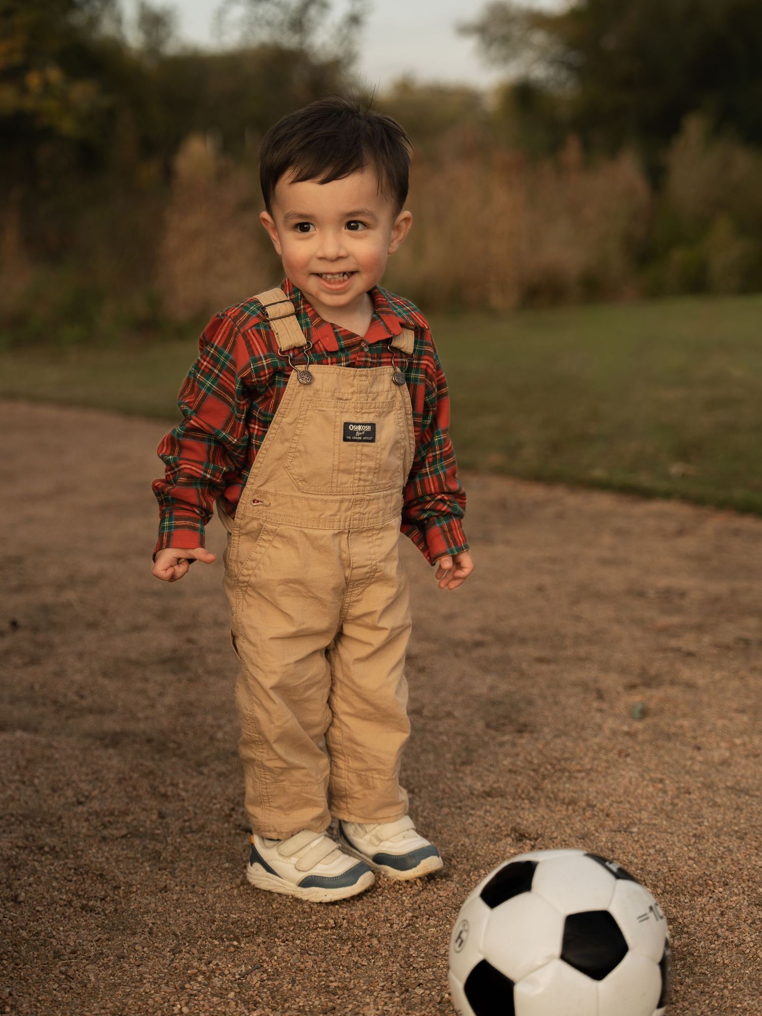 A small child wearing plaid shirt and tan overalls stands on a dirt path next to a soccer ball in an outdoor setting.
