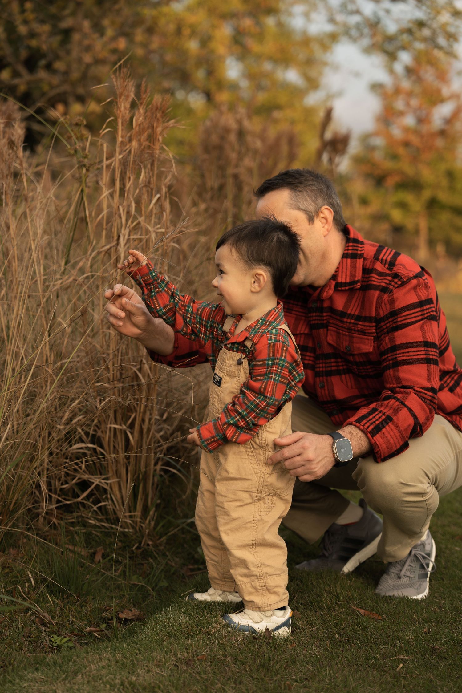 Person wearing red plaid shirt and khaki pants crouches down to explore fall foliage in outdoor nature setting.