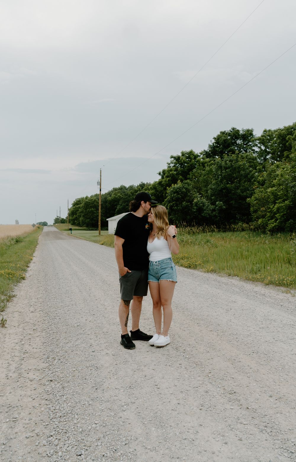 A couple walking together on a gravel country road surrounded by green fields and trees on a summer day.