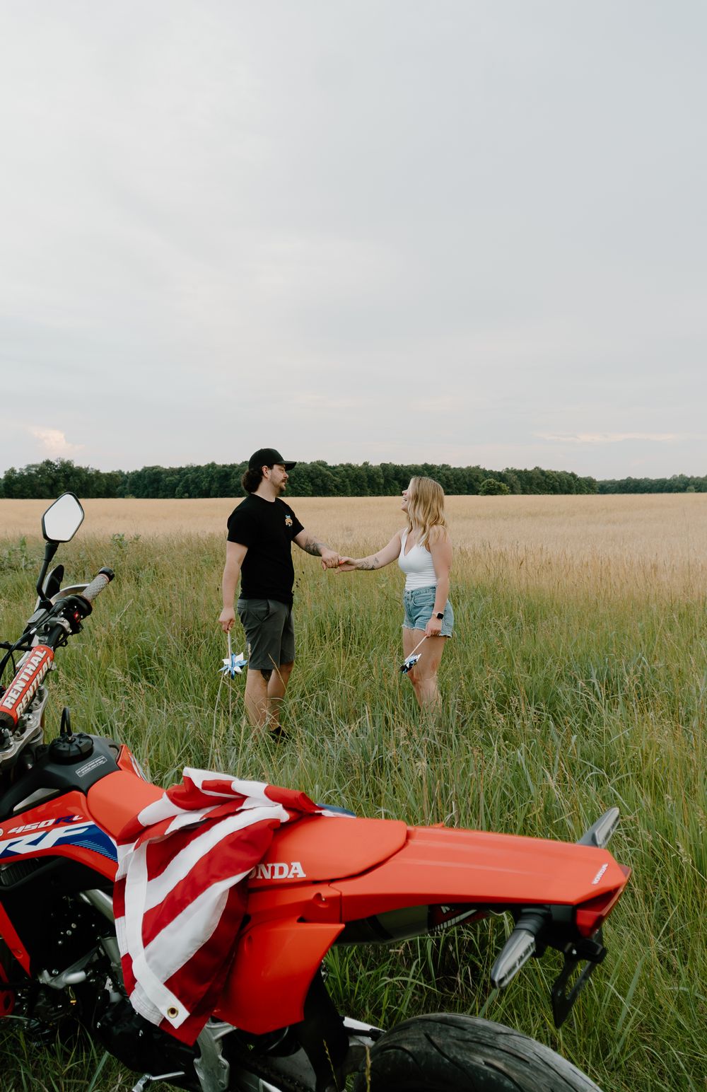 Red Honda motorcycle parked in a tall grass field with two people walking in the distance at sunset.