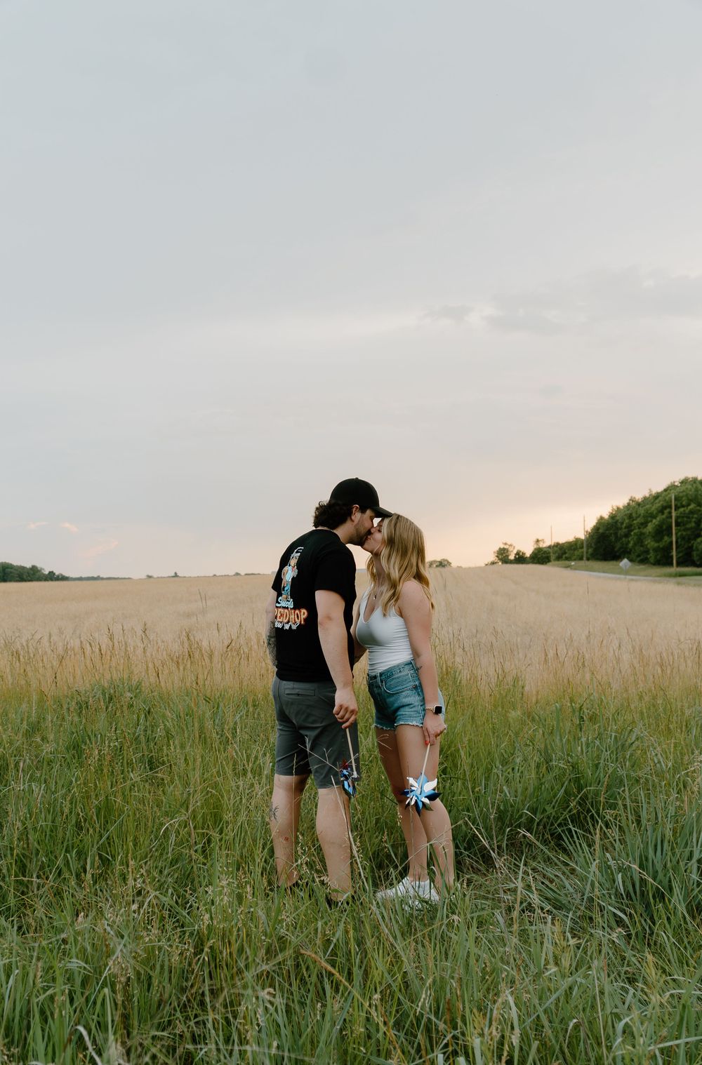 Two people crouch down together in a golden wheat field at sunset with tall grass in the foreground.